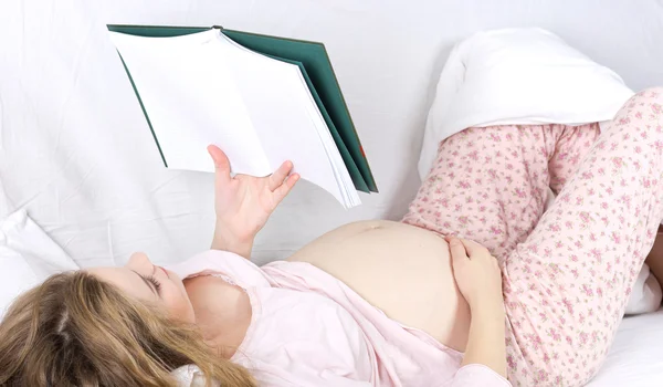 Piękna blondynka w ciąży, czytanie książki w łóżku. Portret kobiety w ciąży — Zdjęcie stockowe