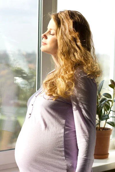 Beautiful pregnant blonde standing near the window and enjoy the sun. — Stock Photo, Image