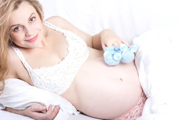 Vackra gravid blond kvinna med tossor i hennes hand.young gravid kvinna luxuriates i en vit säng. Kvinnan Bed Portrait.Beautiful gravid lockiga blonda — Stockfoto