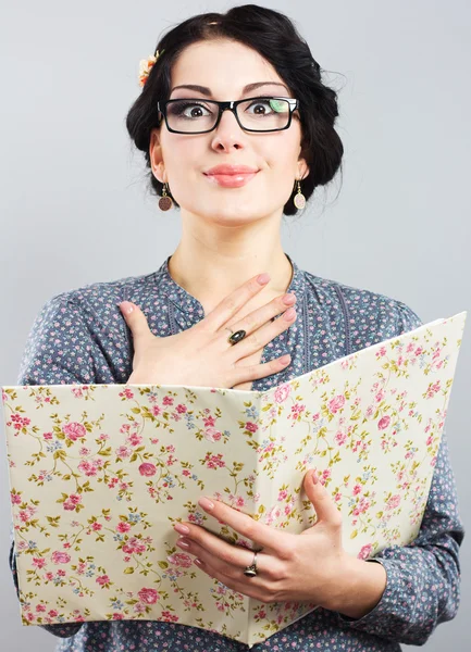 Atractiva estudiante femenina con una carpeta en las manos. Retrato de una hermosa morena sobre un fondo gris. Chica joven con gafas en marco negro —  Fotos de Stock