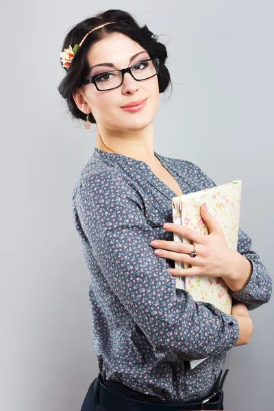 Attractive female student with a folder in hands. Portrait of a beautiful brunette on a gray background. Young girl with glasses in black frame — Stock Photo, Image