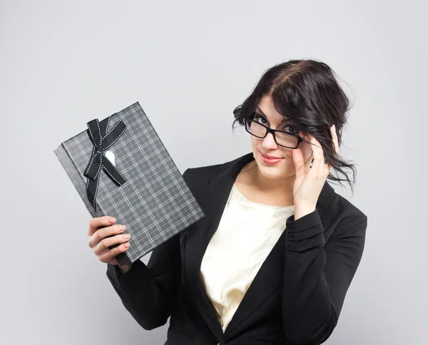 Mujer de negocios en un estricto traje negro con un regalo. Hermosa morena sobre un fondo gris. Regalo . Fotos de stock