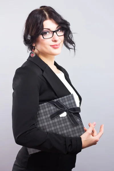 Mujer de negocios en un estricto traje negro con un regalo. Hermosa morena sobre un fondo gris. Regalo . — Foto de Stock