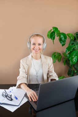 Kulaklıklı, ceketli, gülümseyen genç bir kadın dizüstü bilgisayarın başında oturuyor..