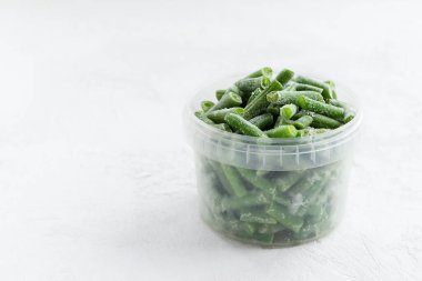 Beyaz bir masada, konteynırda donmuş yeşil fasulye. Yatay yönelim, boşluğu kopyala.