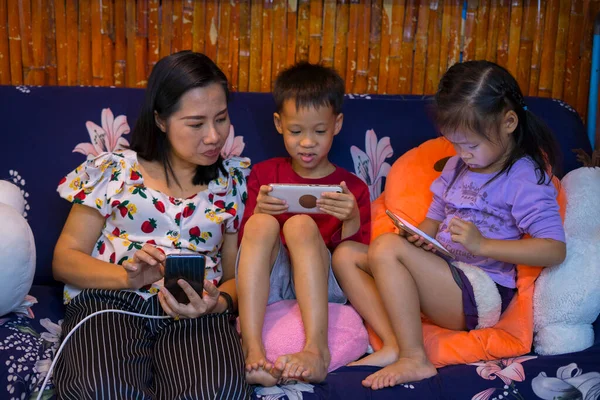 Happy technology addicted Asian family parents and kids use phone sit on couch at home, mom with children obsessed with devices overuse social media, internet addiction concept.