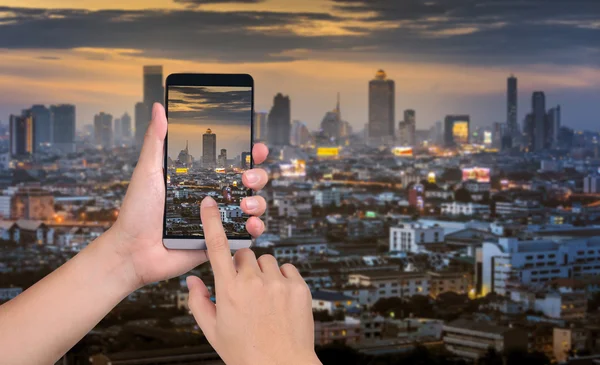 Sostener la mano teléfono inteligente capturar las imágenes en la ciudad puesta del sol de nuevo — Foto de Stock
