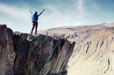 Sırt çantalı bir yürüyüşçü dağın tepesinde duruyor, elleri havada ve güneşin doğuşunun keyfini çıkarıyor.