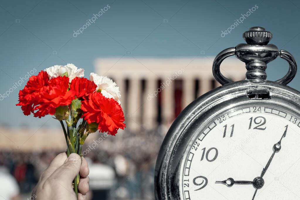Hand holding red and white cloves next to big pocket watch in front of blurry Anitkabir mausoleum in November 10. Old pocket watch showing time 9:05 AM, hour of the death of the leader and founder of Turkish Republic.