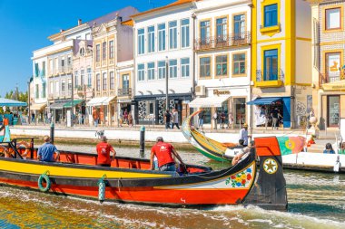 AVEIRO-PORTUGAL: 13 Ekim 2025: İki rehber ve Portekiz bayrağı taşıyan bir moliceiro teknesi Aveiro kanalı boyunca hareket ediyor, tarihi, renkli binalar ve dükkanlardan geçiyor, şehrin hayat dolu cazibesini yakalıyor.