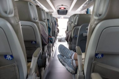 Bir gezgin, koridorda uzanan gri bir otobüs koltuğunun arkasında açık mavi spor ayakkabılarının içinde dinlenir. Tekrarlanan oturma sıraları modern koçun arka planına kadar uzanıyor..
