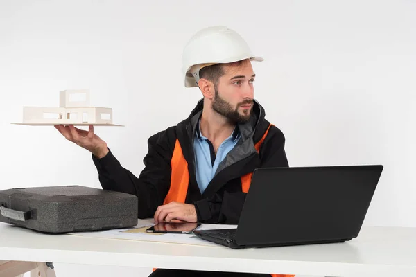 Civil engineer holds a model of the future house on his hand and looks at the person behind the frame. Construction project presentation. Man in a construction uniform sits next to laptop and drawings
