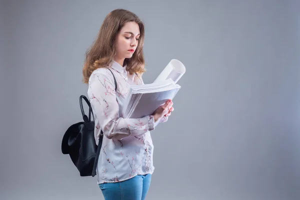 Woman student on gray background. Young girl with a backpack behind her back. The student has a lot of papers in her hands. She\'s thinking about something. Concept - girl student reads thesis.