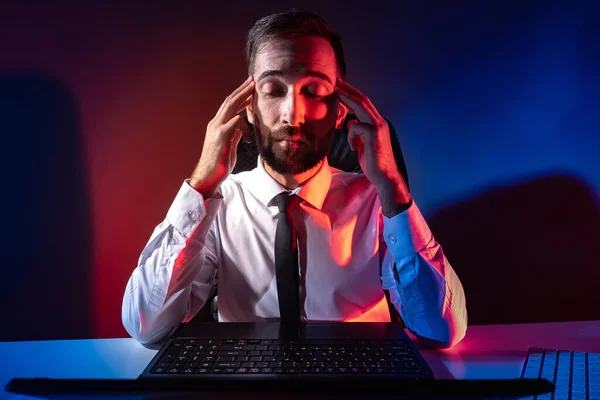 Office worker in a dark office. Man is holding his head. Concept - stress at work. The man next to the laptop. Office worker is under stress. Health problems at work. He has a headache.