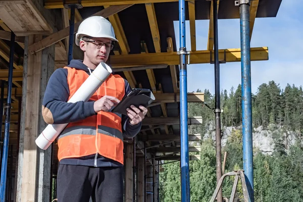 Builder with a tablet and drawings in hands. Builder the background of a house under construction. Concept - architect on a construction site. Man on the background of a building under construction