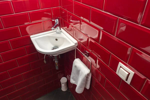 Small sink in WC room. Ceramic sink is fixed in corner. Walls are decorated with red tiles. Towel hangs next to sink. Concept - a place for washing hands in a WC room. Compact washbasin.