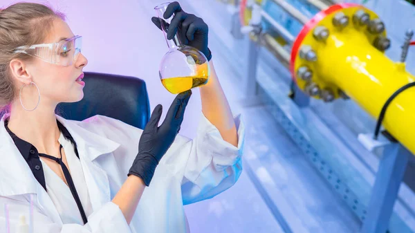 Chemist at the company. Female laboratory assistant examines a yellow chemical liquid in a test tube. Chemist on the background of the pipes of a chemical enterprise. Synthetic detergents production.