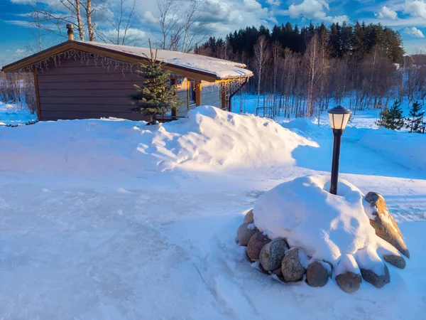 Lake house in winter weather. Wooden house is covered with snowdrifts. Small cottage overlooking forest. Lantern next to a country house. Snow near cottage has been partially cleared.