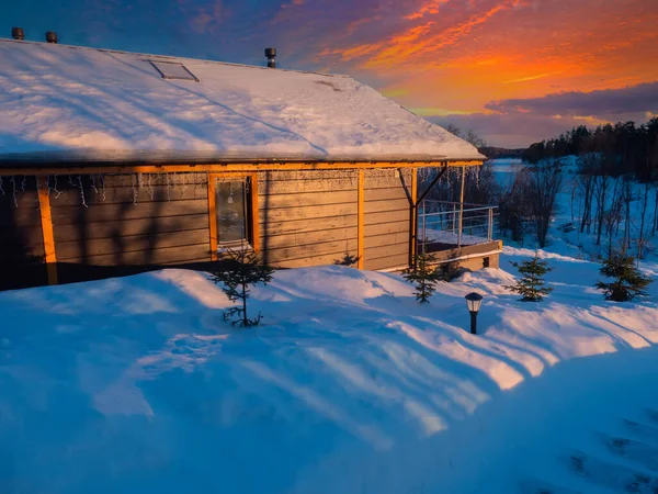 Cottage on a winter day. Private house and snow drifts are illuminated by sunset. Winter sunset. Christmas house on background of a red sunset. One-story cottage overlooking frozen lake. Winter sunset with houses.