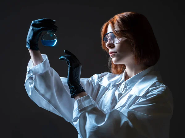 Woman laboratory technician. Young woman Laboratory assistant with a test tube in her hands. Laboratory technician on a dark background. She examines liquid in test tubes. Girl works in medical lab