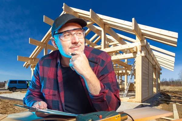 A carpenter working in construction. Carpenter on the background of the frame of a new house. A man in protective glasses on the background of a construction site. A person is engaged in woodworking.