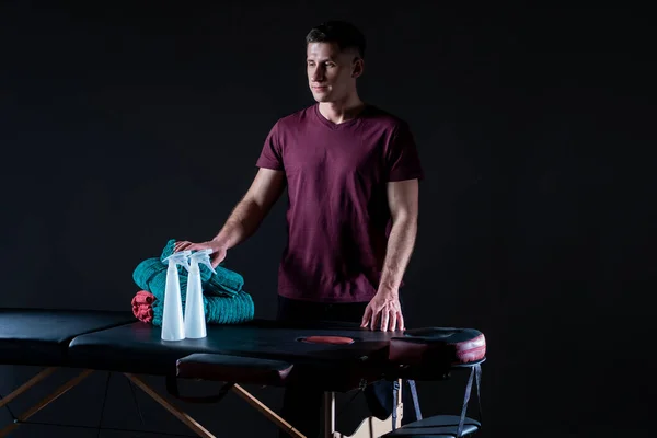 Man works a masseur. He poses next to massage table. Portrait of a male masseur on a black background. Massage therapist stands in dark. Young guy in a massage parlor. He makes a career as a masseur