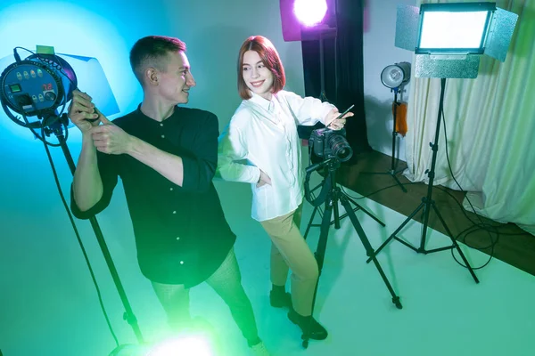 A man and a woman in a photo studio. Lighting and photographic equipment next to them. A photographer sets up equipment for a photo shoot. Concept - services of a professional photographer.