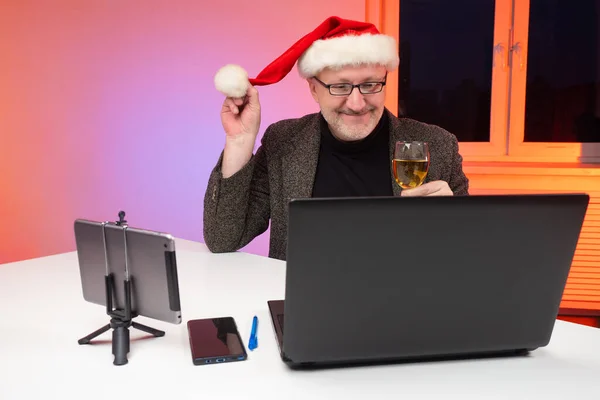 Guy talking to someone via video chat. Laptop and tablet on festive table. He congratulates relatives online. Christmas greetings online. Happy man in a Christmas hat with wine in his hands.