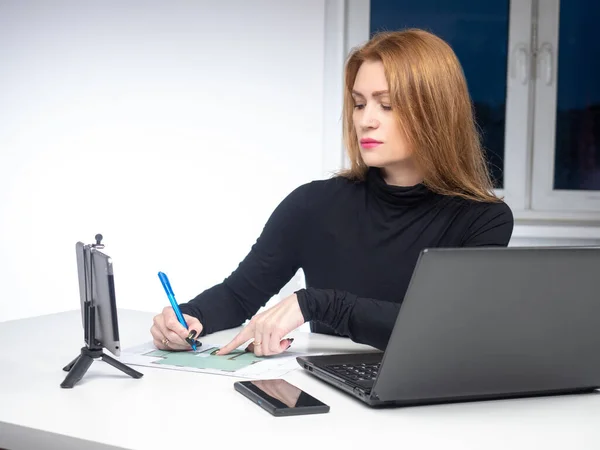 Female architect works remotely. Freelance construction engineer. Remote work on the new building design. Woman is sitting in front of a laptop and tablet. Engineer makes notes in construction plan.