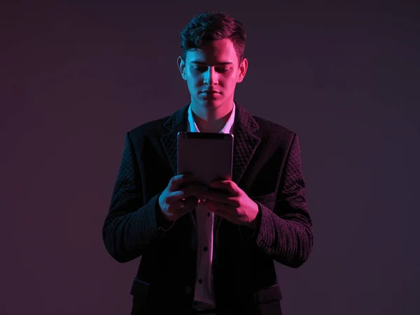 Young businessman with electronic tablet in his hands. Young man in a business suit with a tablet in his hands. Guy with an electronic gadget stands in the middle of the shot on a dark background.