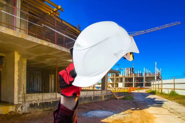 Construction of houses. A hand in a work glove holds a white hard hat against the background of a construction site. Advertising a construction company.