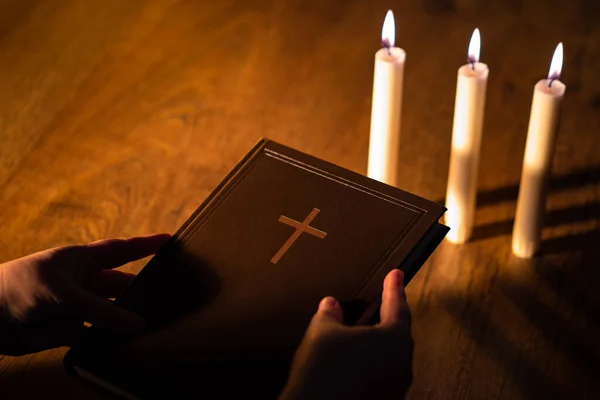 Holy book in hands of a man. Christian Bible. Book with Christian prayers. Concept - reading Christian sermons. Church candles next to hands of a Catholic. Orthodox book with image of cross