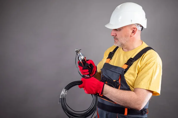 Electrician on a gray background. A man crimps the wires. A worker with a coil of wire and crimpers in his hands. Wiring installation. Working tool. Jobs for workers.