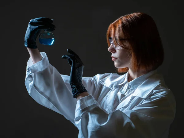 Biological laboratory worker woman. She studies various bacteria. Woman laboratory assistant on a dark background. Lab assistant examines a test tube with some substance. Biologist on dark background