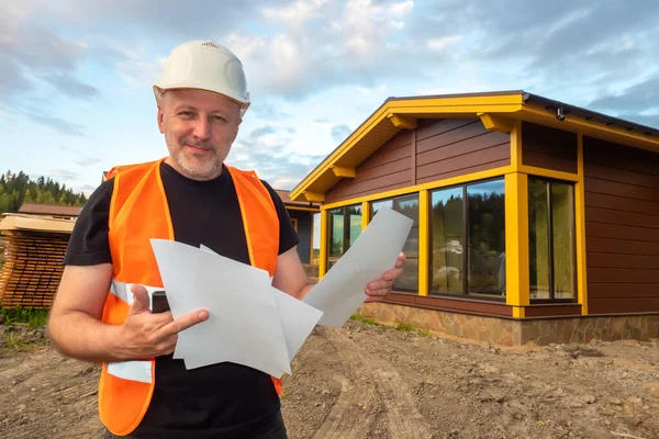 A construction worker in an orange vest smiles at the camera. A man with construction documents and a phone. The engineer stands near the house under construction. Guarantee of work quality.