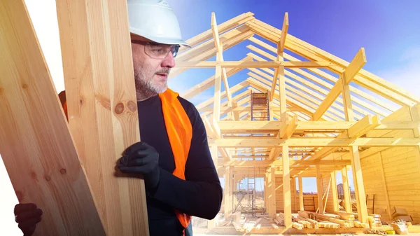 Construction of cottages. A man with boards on the background of the frame of the future house. A construction worker in a white hard hat and orange vest. Work of a construction company.