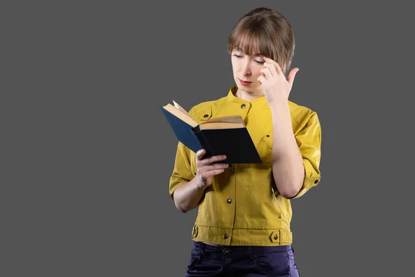 Female student reads a book. Concept - she is preparing for the exam. Young woman with a book in her hands. Girl student with a textbook. A woman in a yellow jacket is holding a book.