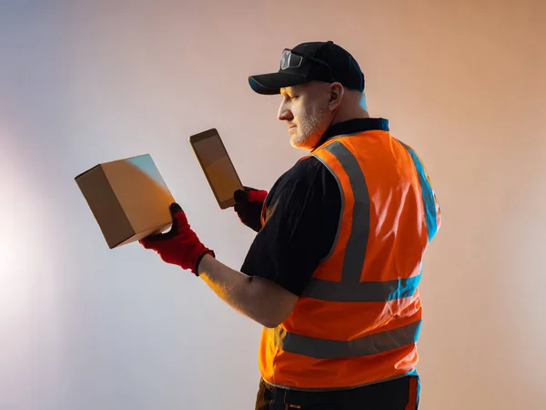 Warehouse manager in an orange vest. Warehouse manager at fulfillment center. Warehouse worker with a cardboard box in his hands. Man with electronic tablet. Man on light background.