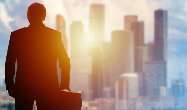 Business start. A man in a business suit looks at the business center of the city. Person and the skyscrapers are illuminated by the rising sun. A man with a briefcase in his hands.