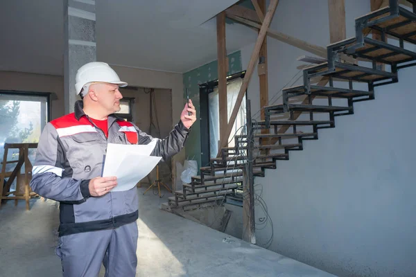 Builder is talking via video link. Repairman holds phone in front of him. Man in a gray uniform. Concept - he makes a video report for customer. Workplace builder. Room construction in background