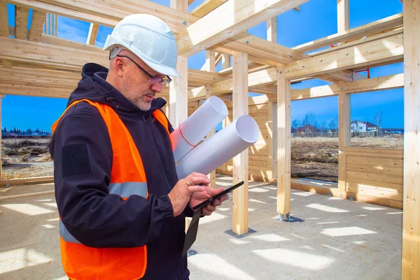 The estimate of construction. The cost of a new home. A man with drawings and a mobile phone on the background of a cottage under construction. A Builder in a white helmet on a construction site.