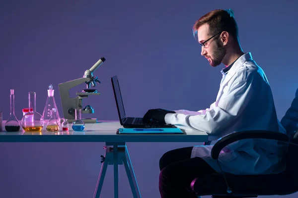 Medical researcher next to laptop. Medic works in a dark lab. Concept - it conducts medical researc. Test tubes and a microscope on doctor table. Researcher works in a laboratory. Doctor in lab