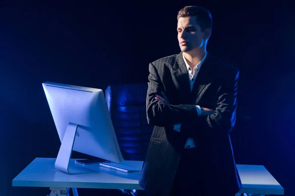 Gloomy businessman next to computer. Man in a business suit near to office table. Young businessman stands with his arms crossed. He not happy about something. Guy crossed his arms standing in office