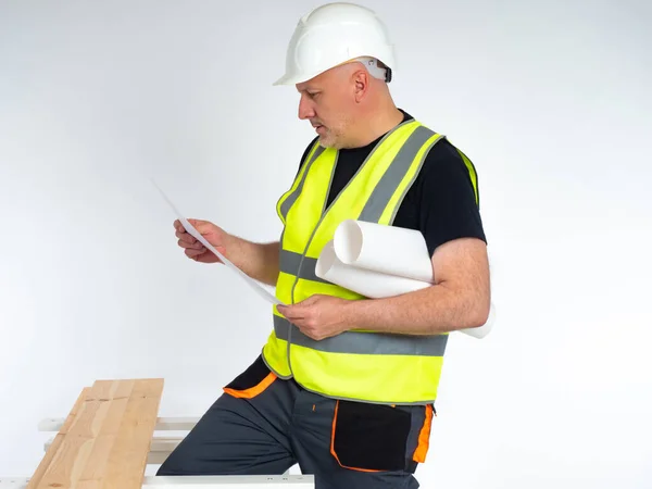 Carpenter in a yellow vest and hard hat. Man next to the boards. The carpenter examines a sheet of paper. Concept - a carpenter at a construction site. Builder on a white background.