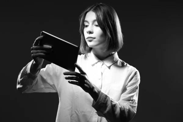 Girl with a tablet in black and white. The girl is watching a video on a tablet PC. A teenager with a computer gadget. The girl is playing on a tablet. Computer games.