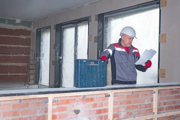 Engineer in a building under construction. A man in a uniform and a white helmet studies the plans of a building under construction. Construction and decoration of the office.