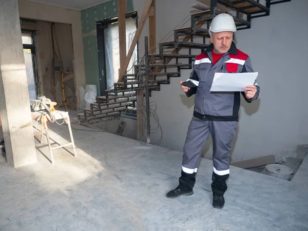 Repairman inside a house under construction. Repairman in uniform with blueprints in his hands. Man in a construction worker uniform. Career as apartment renovator. He examines a sheet of paper