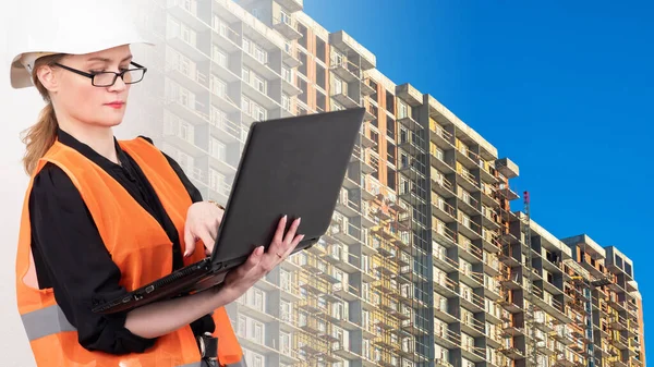 Woman works in construction industry. Woman builder with a laptop in her hands. Woman on background of a building under construction. Girl in uniform of a builder. Construction of high-rise buildings