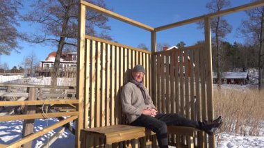 Trosa, İsveç Kış güneşinde bir adam donmuş bir gölün kıyısında, ahşap bir güneş güvertesinde oturur.