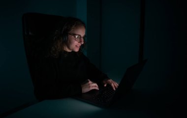 Geceleri dizüstü bilgisayar kullanan, kablosuz kulaklıklı, gülümseyen kadın. Öğrenci geç saate kadar çalışıyor.